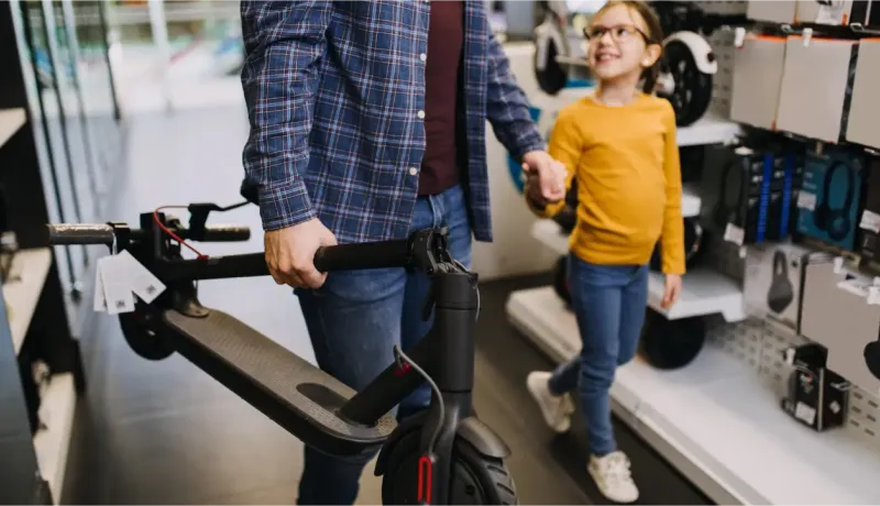 Un homme tient une trottinette électrique tandis qu'une petite fille à lunettes, souriante, lui tient la main dans un magasin.