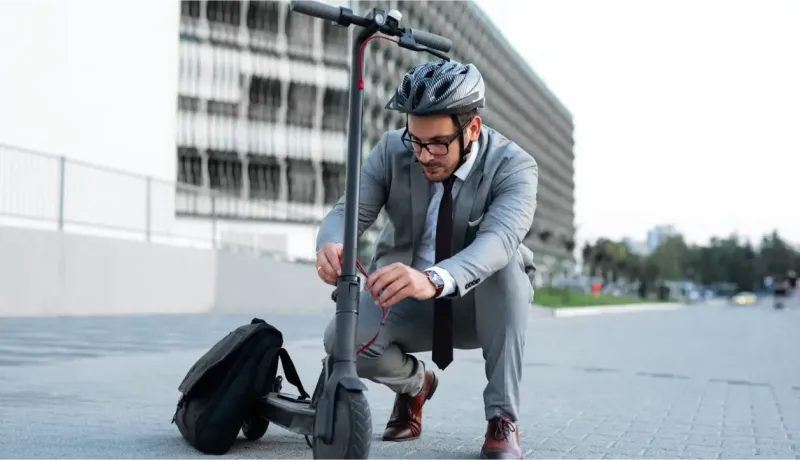 Un homme en costume et casque ajuste sa trottinette électrique dans un environnement urbain moderne.