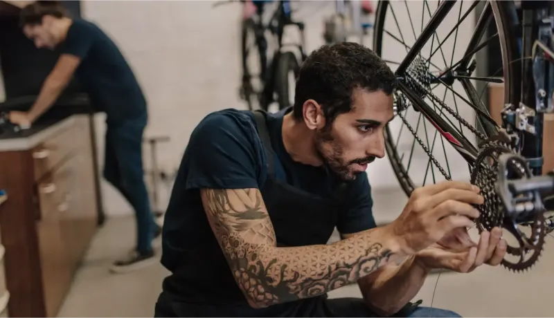 Un mécanicien barbu avec des bras tatoués répare la chaîne d'un vélo dans un atelier, un autre homme en arrière-plan.