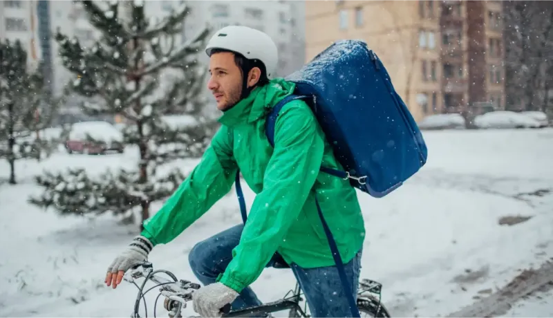 Un livreur à vélo avec un casque blanc et un grand sac bleu pédale sous la neige.