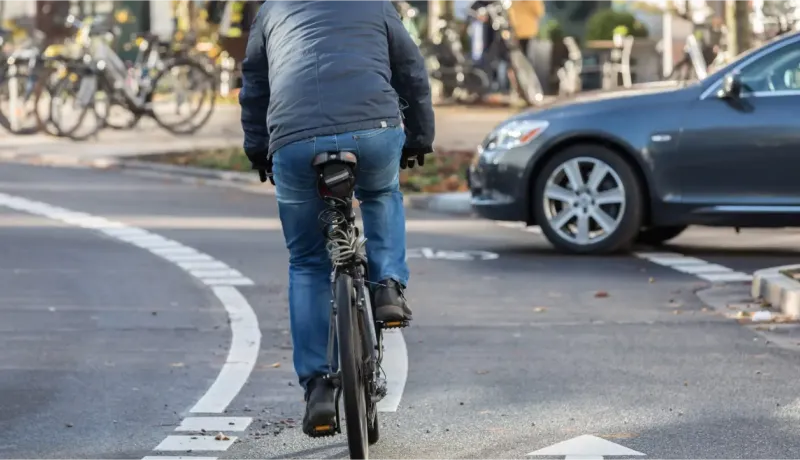 Un cycliste en doudoune et jean bleu se déplace sur une piste cyclable. On peut voir la roue arrière de son vélo et l'arrière d'une voiture.