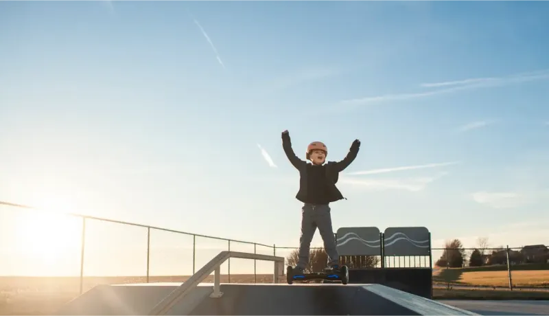 Un enfant casqué lève les bras en signe de victoire sur un hoverboard, dans un skatepark sous un ciel bleu.