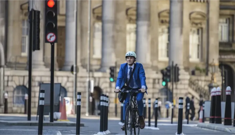 Un homme en costume et casque roule à vélo sur une rue bordée de bâtiments classiques, avec un feu rouge en arrière-plan.