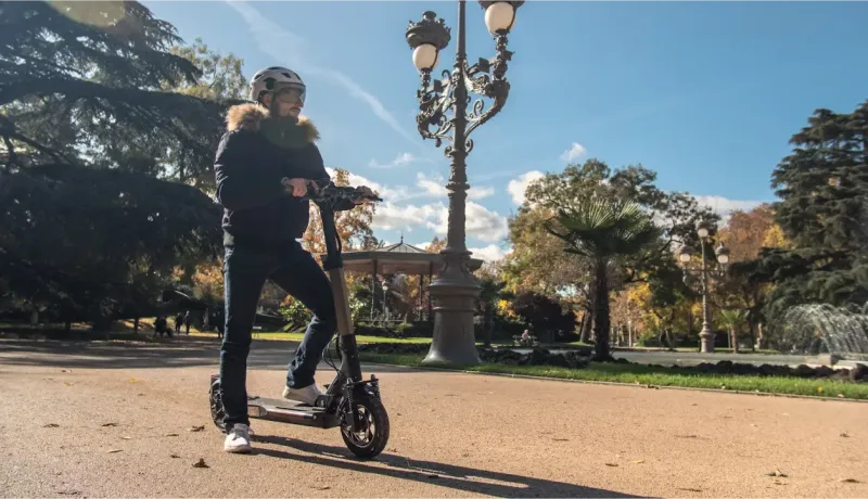 Un homme en casque et veste d'hiver se tient à côté d'une trottinette électrique dans un parc ensoleillé.