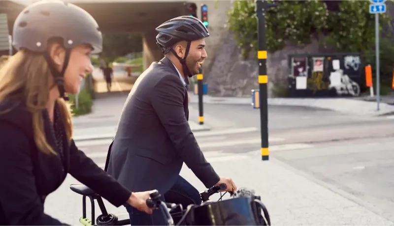 Deux personnes souriantes, casquées et en tenue de ville, font du vélo en milieu urbain.