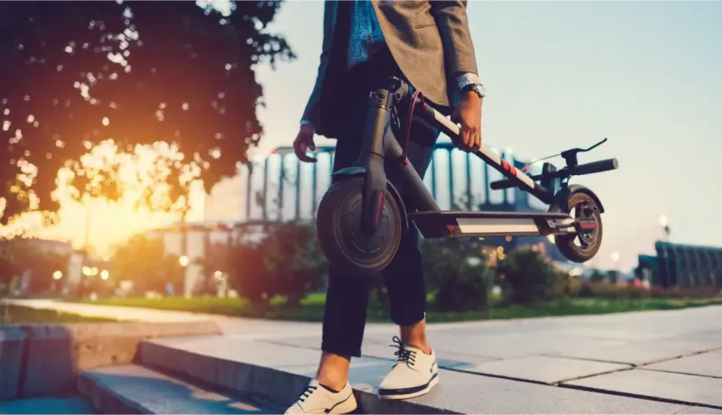 Une personne tenant une trottinette électrique pliée, marchant sur des escaliers extérieurs au coucher du soleil.