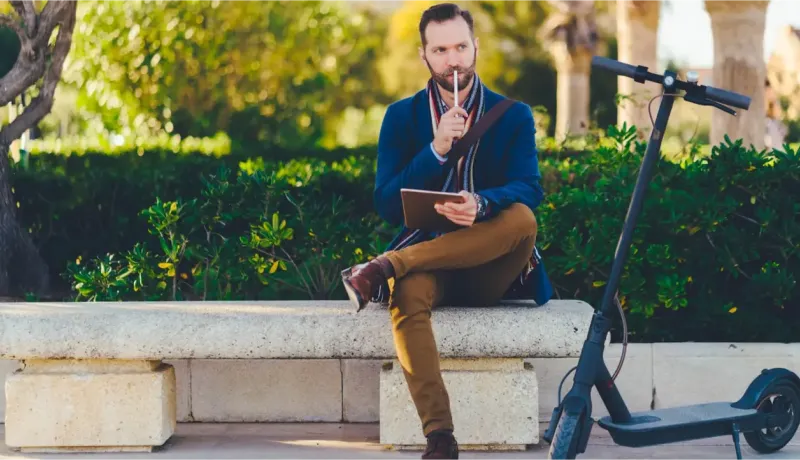 Un homme barbu assis sur un banc, pensif, avec sa trottinette électrique noire garée à côté de lui.