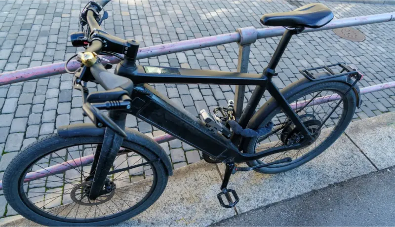 Un vélo électrique noir, avec un antivol attaché, est garé contre une barrière dans une rue pavée.