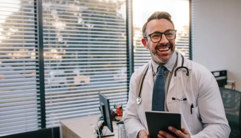 Médecin souriant avec une tablette dans son bureau ensoleillé.