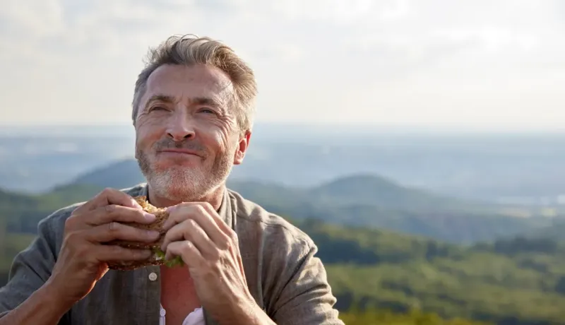 Un homme souriant déguste un sandwich en plein air, avec une vue panoramique sur des collines verdoyantes sous un ciel légèrement nuageux.