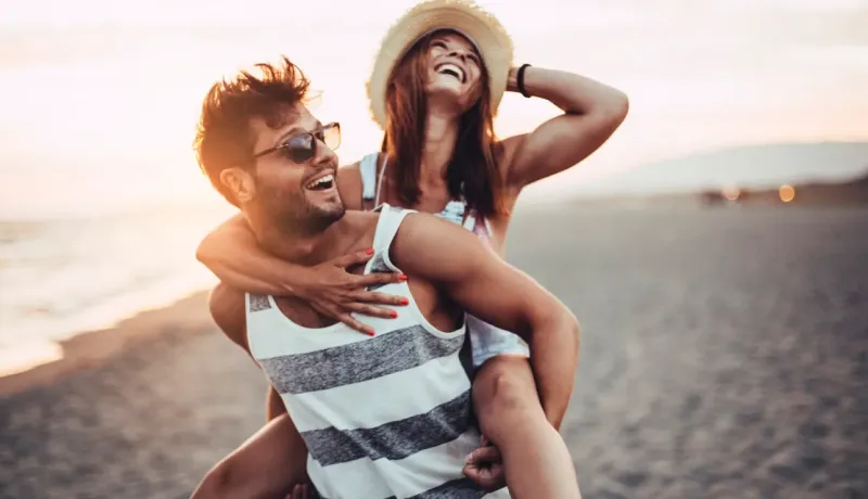 Un couple joyeux s'amuse sur la plage au coucher du soleil, la femme portant un chapeau de paille et riant sur le dos de l'homme.