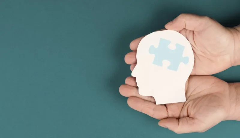 Deux mains tiennent une silhouette de tête en papier avec une pièce de puzzle bleue, symbolisant la santé mentale et la réflexion.