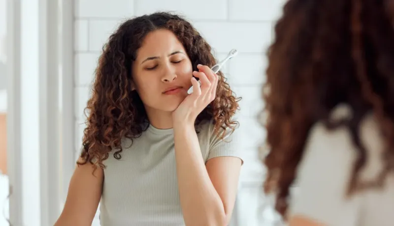 Une femme aux cheveux bouclés ressent une douleur dentaire, tenant une brosse à dents, dans une salle de bain lumineuse.