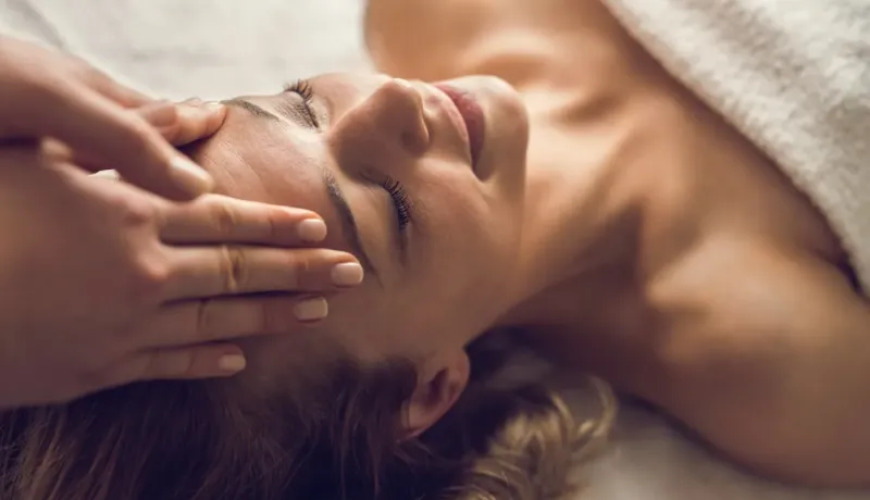 Une personne reçoit un massage relaxant du visage, allongée sous une serviette blanche, les yeux fermés et un sourire apaisé.