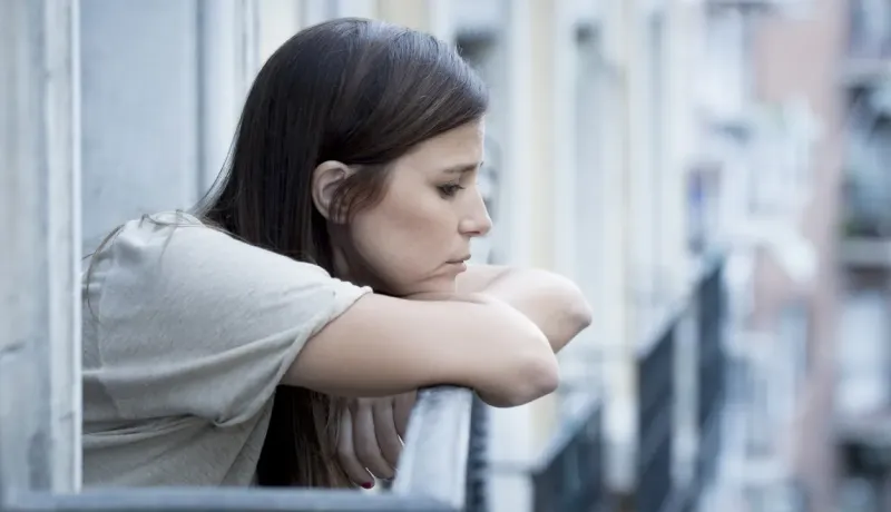 Une femme pensive appuyée sur une rambarde de balcon, regardant au loin, entourée d'immeubles flous en arrière-plan.