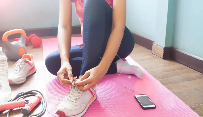 Personne attachant ses lacets sur un tapis de yoga rose, entourée d'équipements de sport et d'un smartphone.