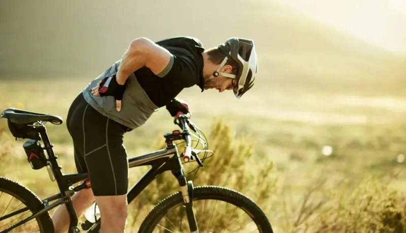 Un cycliste en tenue de sport s'appuie sur son vélo, se tenant le dos, dans un paysage ensoleillé et naturel.