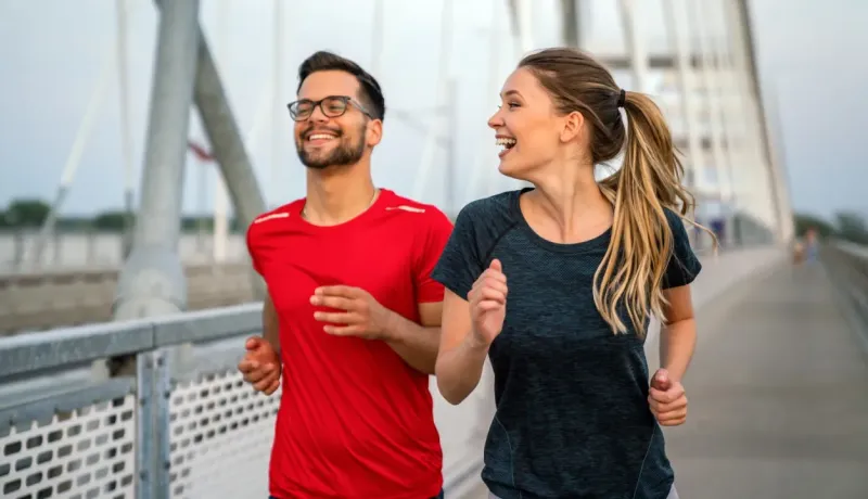 Un homme et une femme courent joyeusement sur un pont, vêtus de tenues de sport colorées, profitant d'une séance d'exercice en plein air.