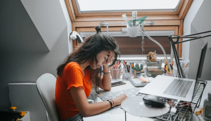 Une adolescente assise à un bureau encombré, la tête appuyée sur sa main, semble stressée ou fatiguée en travaillant sur un ordinateur portable.