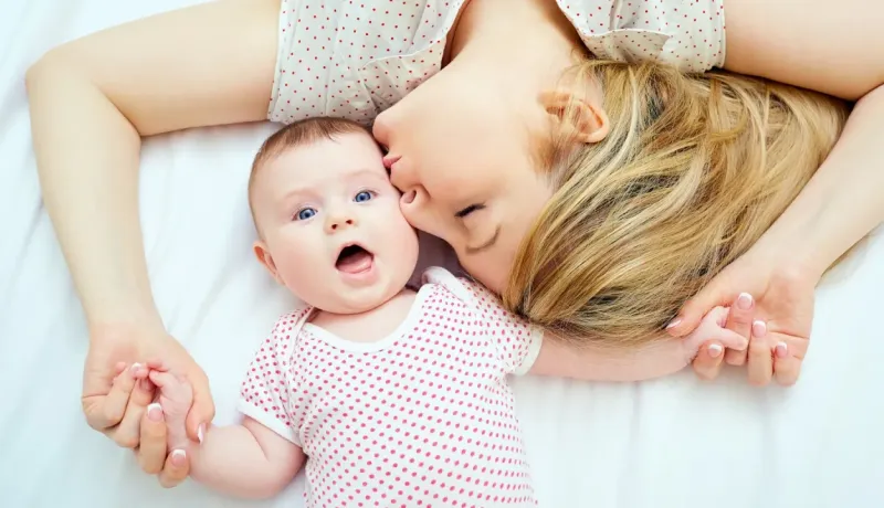 Une femme allongée sur le dos embrasse tendrement un bébé aux yeux bleus sur un lit blanc.