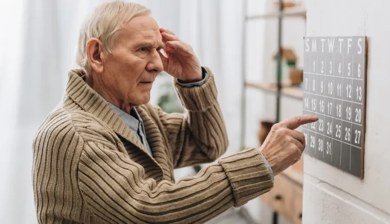 Un homme âgé en pull beige consulte un calendrier mural, pointant une date avec attention, dans un intérieur lumineux et moderne.