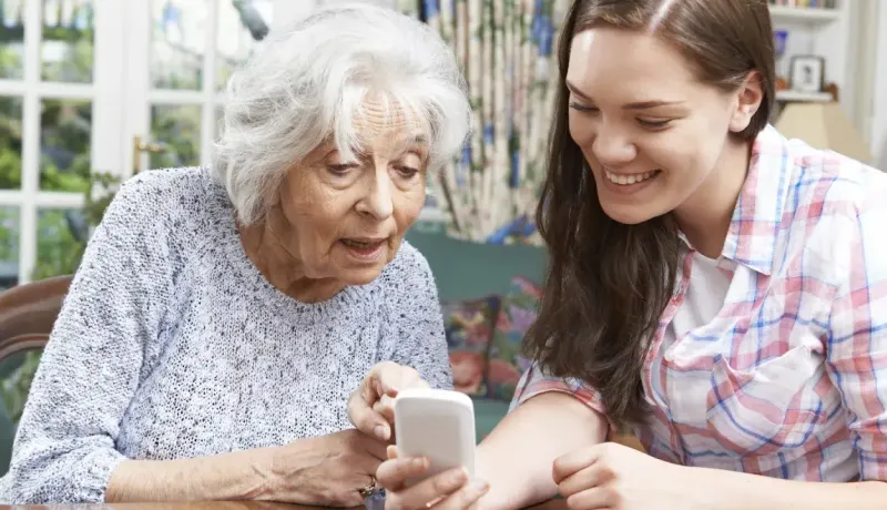 Une jeune femme souriante montre quelque chose sur un smartphone à une femme âgée, assise ensemble dans un salon lumineux.
