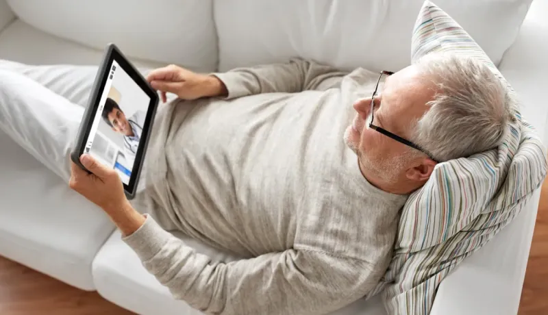 Un homme âgé se détend sur un canapé tout en consultant un médecin sur tablette.