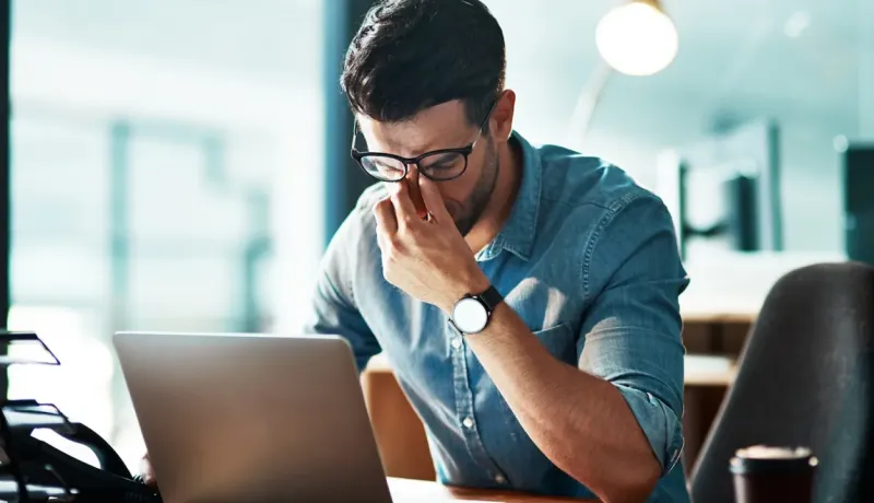 Un homme se frotte les yeux devant son ordinateur, signe de stress/fatigue.