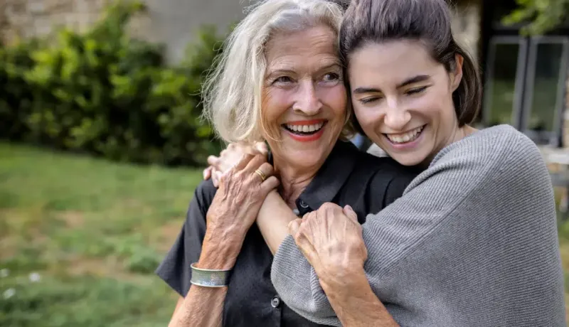Une jeune femme serre dans ses bras une femme âgée souriante à l'extérieur.