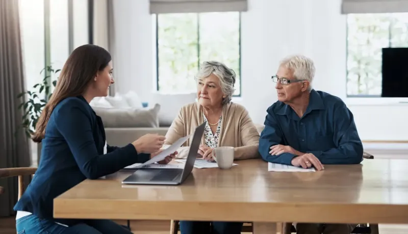 Un couple âgé consulte une conseillère à leur domicile, documents et ordinateur sur la table.