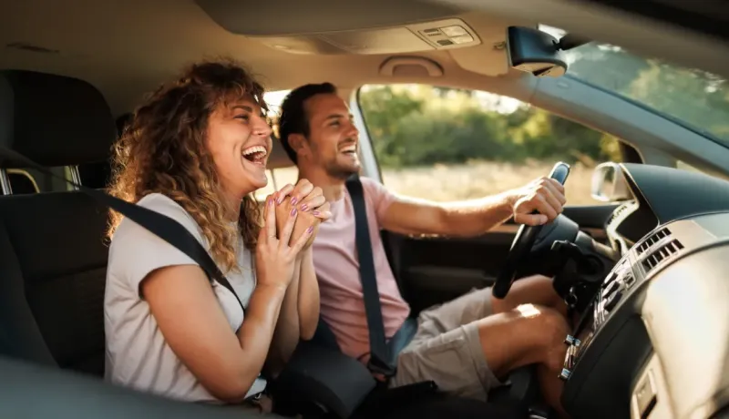 Un couple joyeux et rieur à l'intérieur d'une voiture, la femme en passagère et l'homme au volant, profitant du trajet.