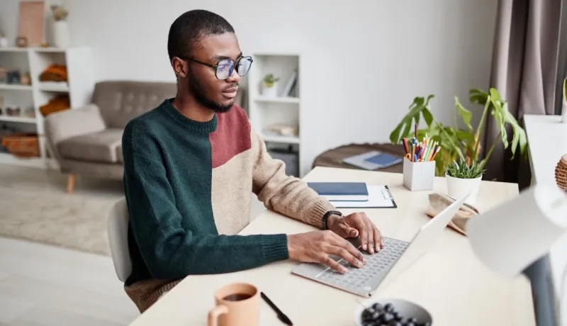 Jeune homme en pull coloré et lunettes, travaillant sur son PC portable sur un bureau clair à la maison. Télétravail.