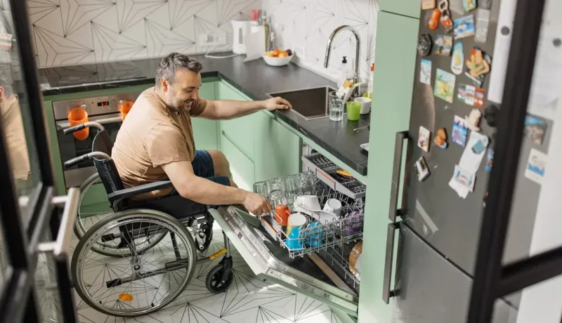 Un homme en fauteuil roulant sourit en rangeant la vaisselle dans le lave-vaisselle de sa cuisine verte et moderne.