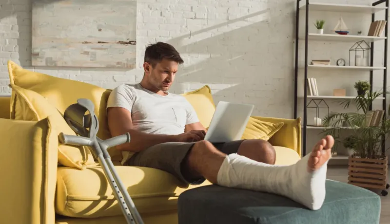 Un homme à la jambe plâtrée utilise son ordinateur portable, confortablement installé sur un canapé jaune avec des béquilles.