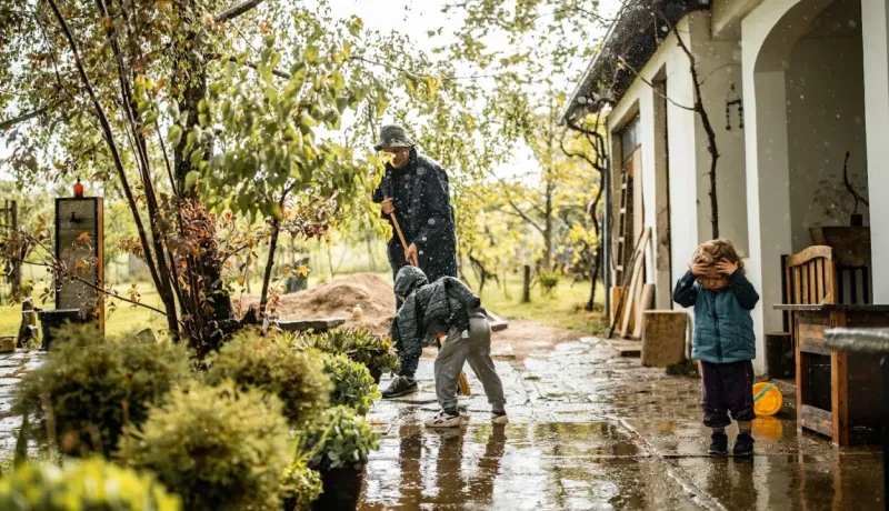 Jour de pluie. Un grand-parent et deux enfants travaillent ou jouent dans un jardin. Scène extérieure pluvieuse.