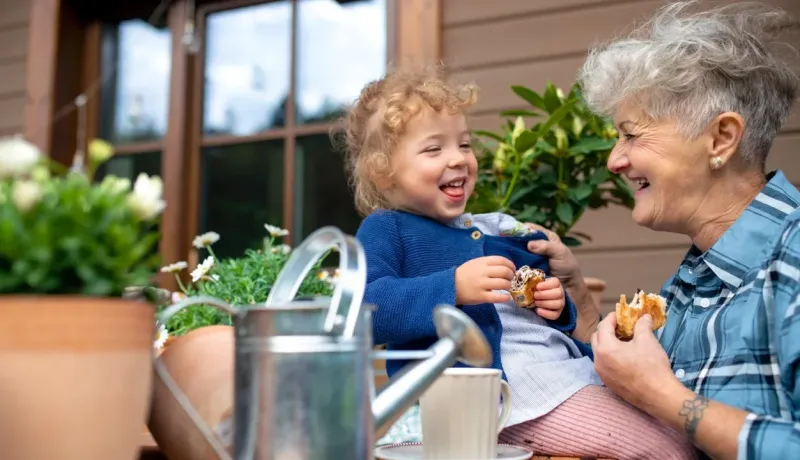 Une grand-mère et un jeune enfant rient ensemble sur un patio, partageant une collation. Scène de famille joyeuse.