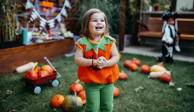 Petite fille souriante, déguisée en citrouille, debout dans l'herbe au milieu de vraies citrouilles pour Halloween.