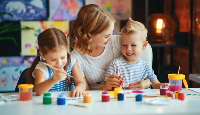 Une mère et ses deux jeunes enfants peignent ensemble à table. Moment de créativité et de joie en famille.