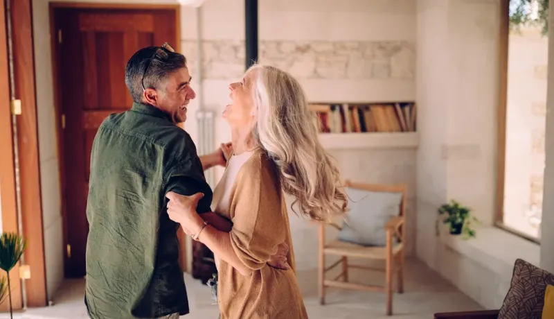 Un couple de seniors joyeux et élégant danse dans son salon lumineux. Moment de bonheur et de complicité.