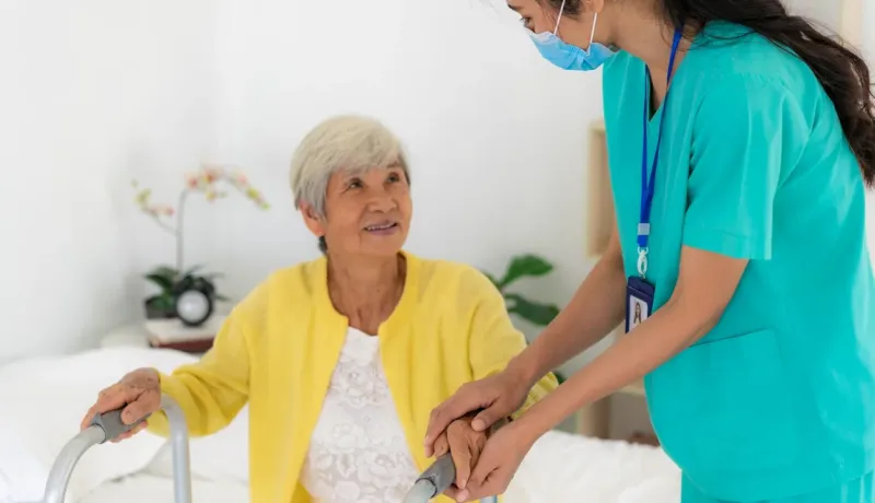 Une infirmière masquée en tenue verte aide une femme âgée souriante à utiliser son déambulateur. Soins à domicile.