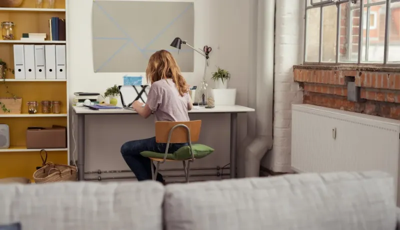 Une femme aux cheveux blonds travaille sur un PC portable à un bureau, dos à la caméra, dans un loft lumineux.