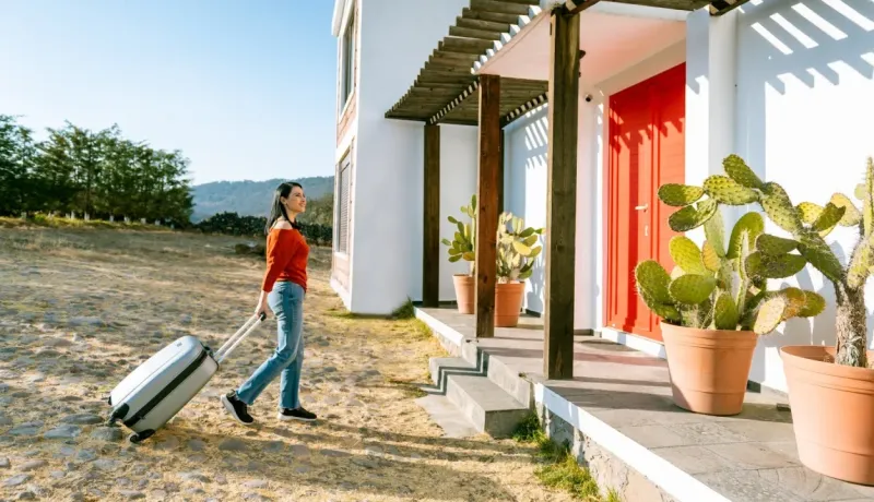 Une femme tire une valise à roulettes vers l'entrée d'une maison moderne, entourée de cactus. Départ en vacances.