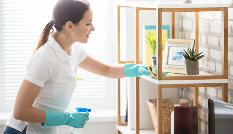 Une femme portant des gants bleus dépoussière une étagère en bois avec un spray.