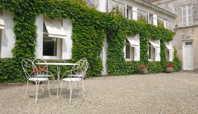 Maison de campagne recouverte de lierre, avec des volets blancs et une table de jardin en fer forgé sur graviers.