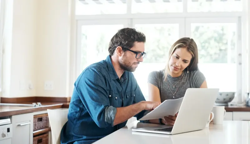 Un couple discute de documents (financiers ou immobiliers) devant un ordinateur portable dans leur cuisine lumineuse.