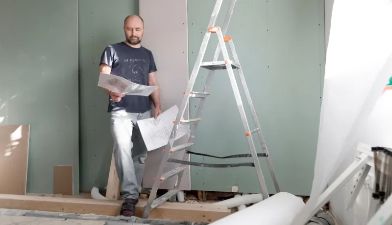Un homme (entrepreneur/ouvrier) examine des plans de construction ou de rénovation devant un mur de placoplâtre.