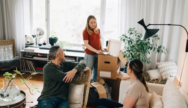 Une famille (mère, père, fille) est dans son salon, déballant des cartons lors d'un déménagement. Ambiance de foyer.