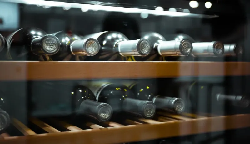 Gros plan de plusieurs bouteilles de vin stockées horizontalement sur des étagères en bois, à l'intérieur d'une cave à vin éclairée.