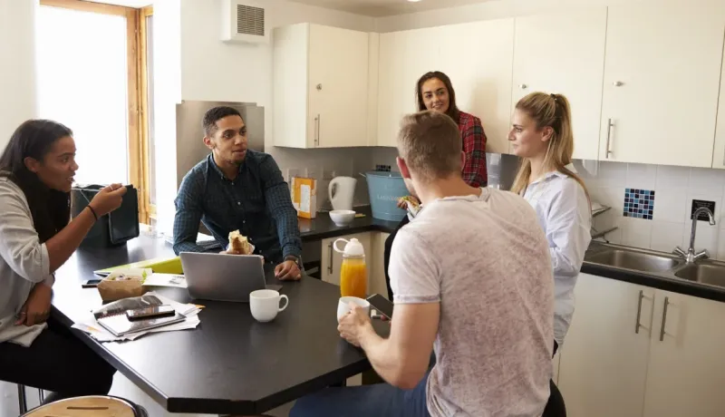 Cinq étudiants ou jeunes collègues discutent et travaillent autour d'une table noire dans une cuisine moderne et lumineuse.