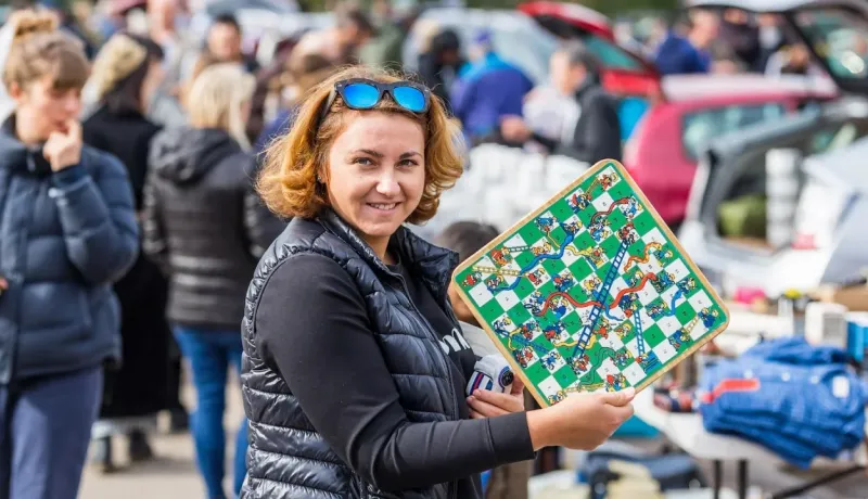 Femme souriante tenant un petit jeu de l'oie ou échelles et serpents lors d'une brocante ou d'un vide-grenier.
